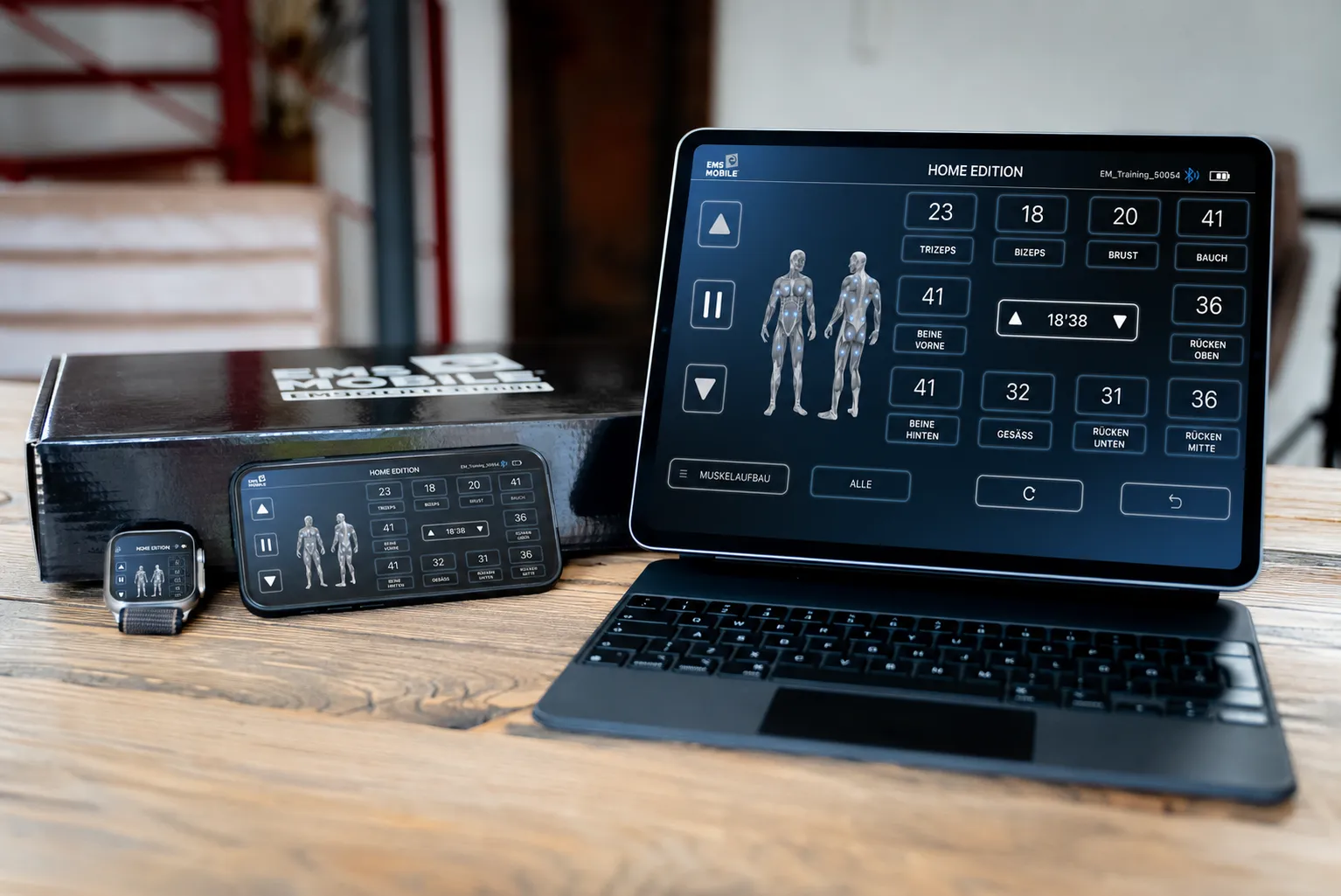 Apple Watch, iPhone und iPad zeigen die EMS Mobile Home-Steuerung mit Muskelgruppen-Übersicht — auf einem Holztisch neben der EMS-Mobile-Box.